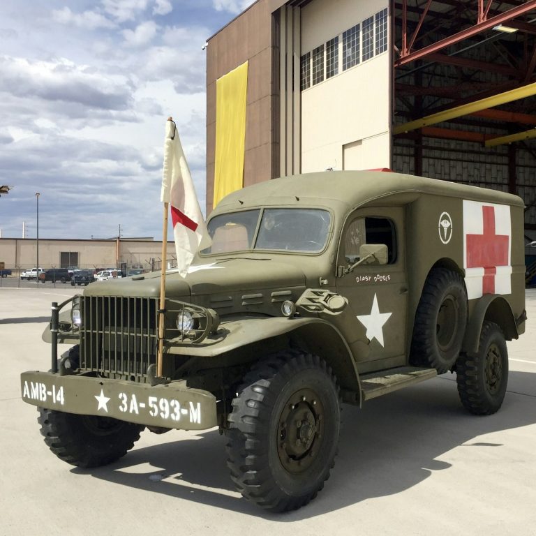 Ein grüner Militärkrankenwagen mit rotem Kreuz steht vor einem Hangar.
